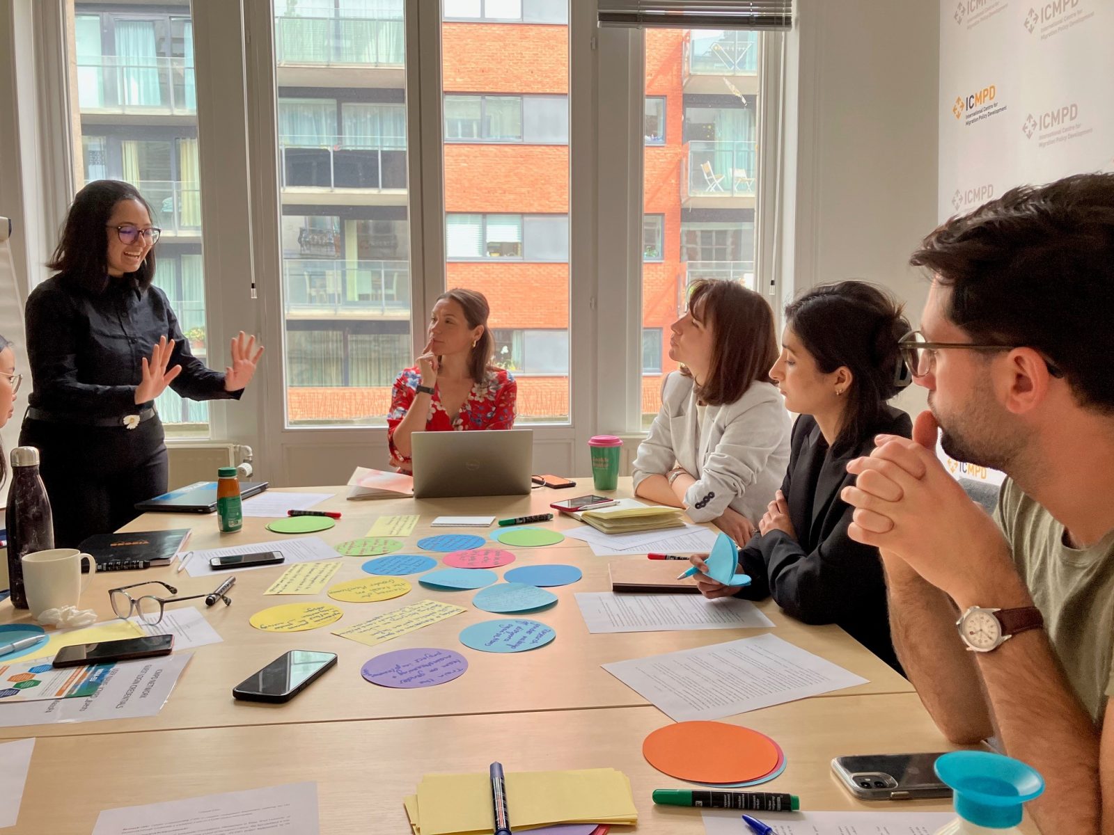 A young woman who is standing up is speaking animatedly to a group of people around a table covered with colourful paper and notes.