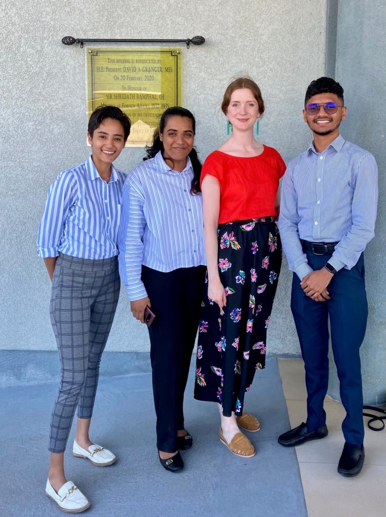 Three smartly dressed women and a man stand together in front of a government building. They are smiling at the camera.