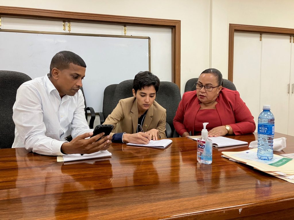 A man and two women at a table. One women is writing in a notebook as the other two look on.