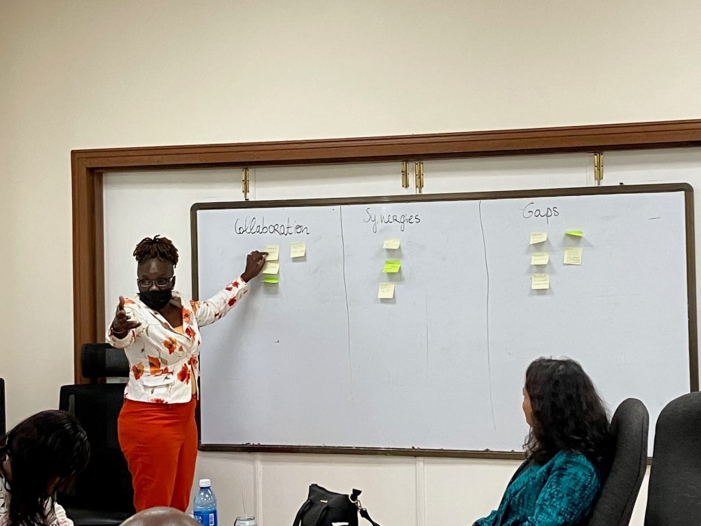 A woman in front of a white board which has sticky notes on it. She is pointing at one sticky note and gesturing towards an unseen audience.
