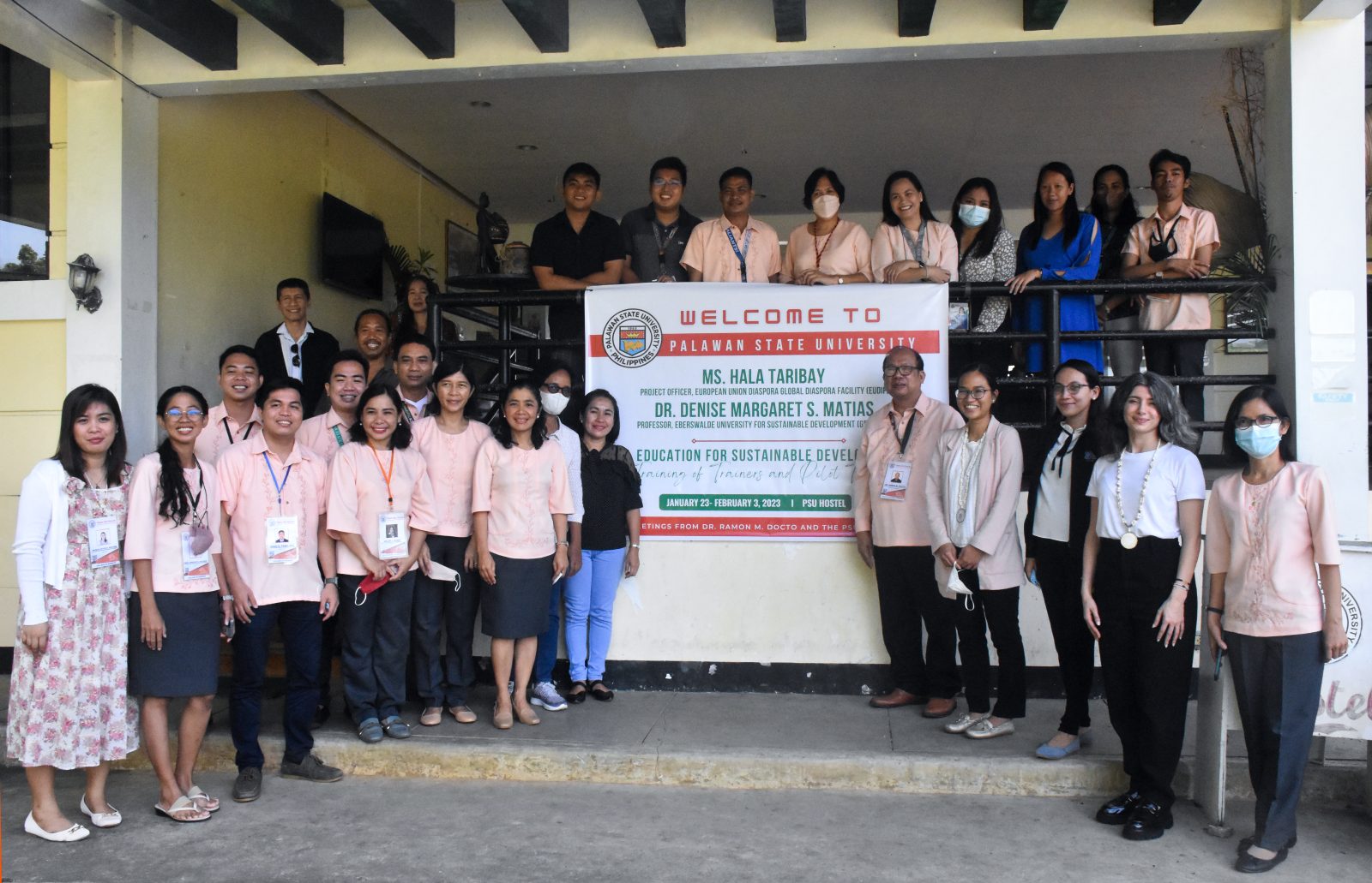 A group of teachers stand in front of a school building on which there is a sign welcoming two visitors for a workshop.