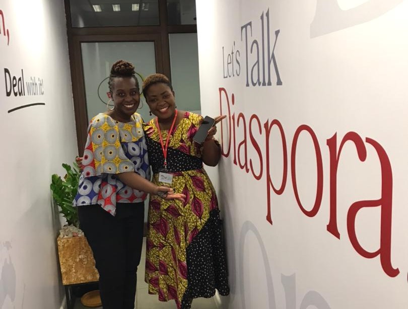 Two smiling women point at the writing on the wall next to them which reads "Let's talk diaspora."